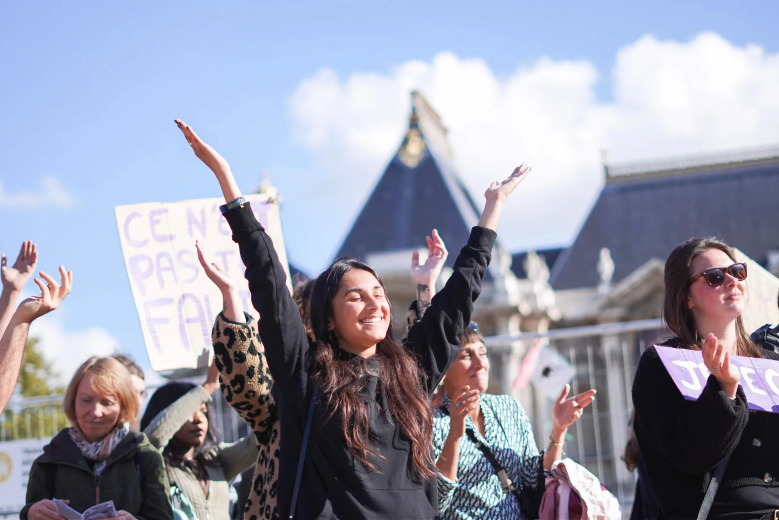 Une femme souriante dans une manifestation, d'autres femmes l'entourent. Derrière elle, une pancarte surexposée, et difficile à lire, sur laquelle on peut devinier "Ce n'est pas ta faute"